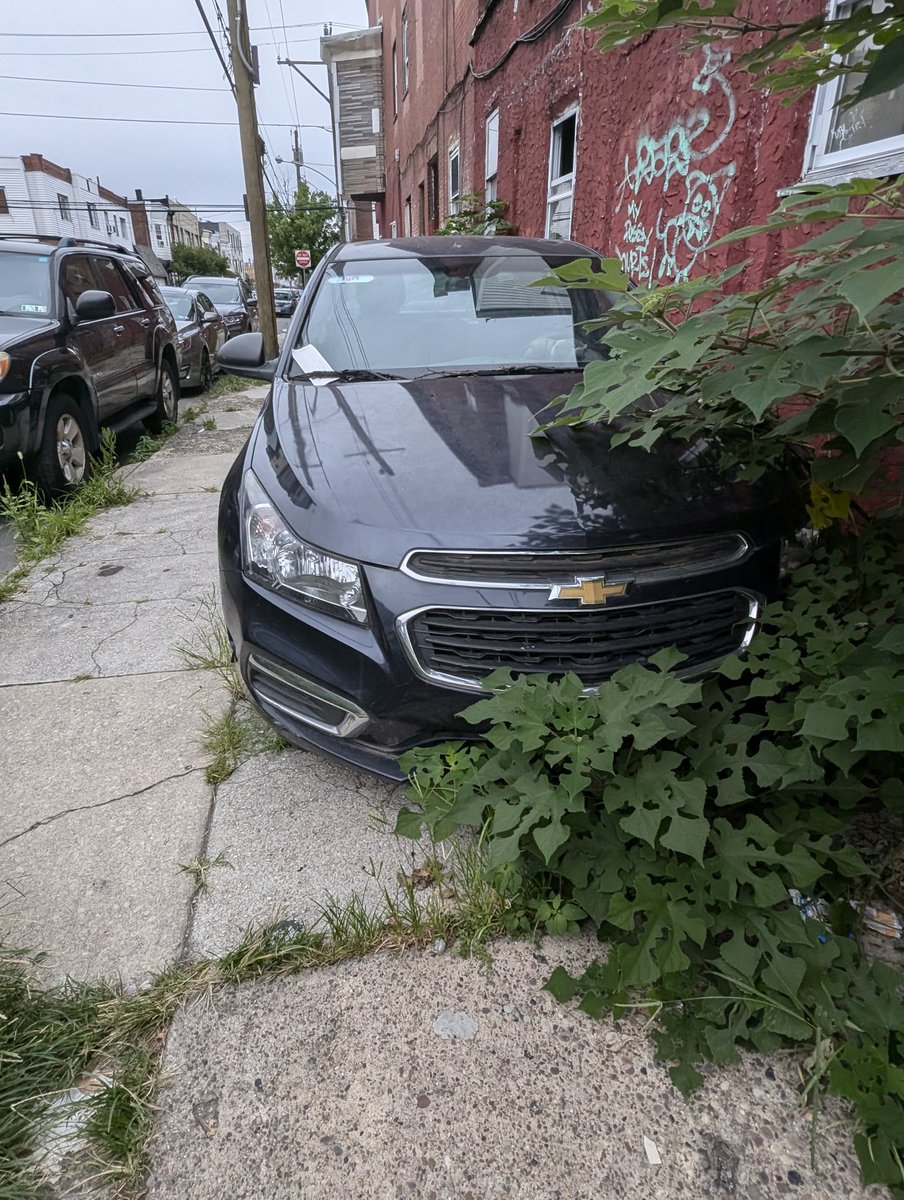 A car parked on the sidewalk next to an abandoned building. Weeds grow up around it.