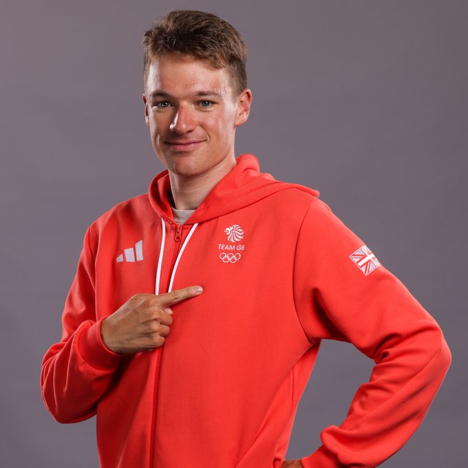 Team GB cyclist Ethan Hayter stands in a red hoodie and points with his right hand across his chest to the Team GB logo