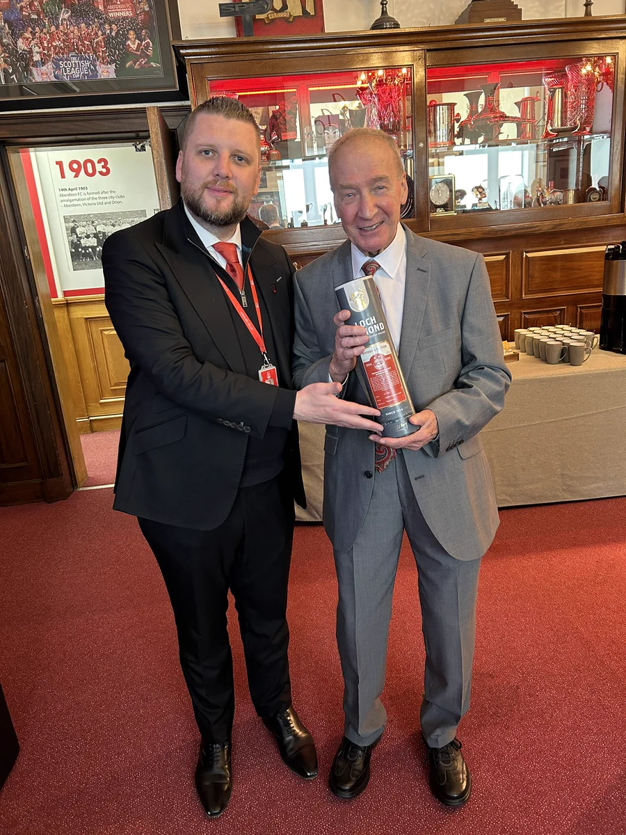 Alan Burrows on left of photo wearing black suit presenting bottle of whisky to John wearing grey suit. In the boardroom, red carpet on floor with glass fronted display cabinets behind them