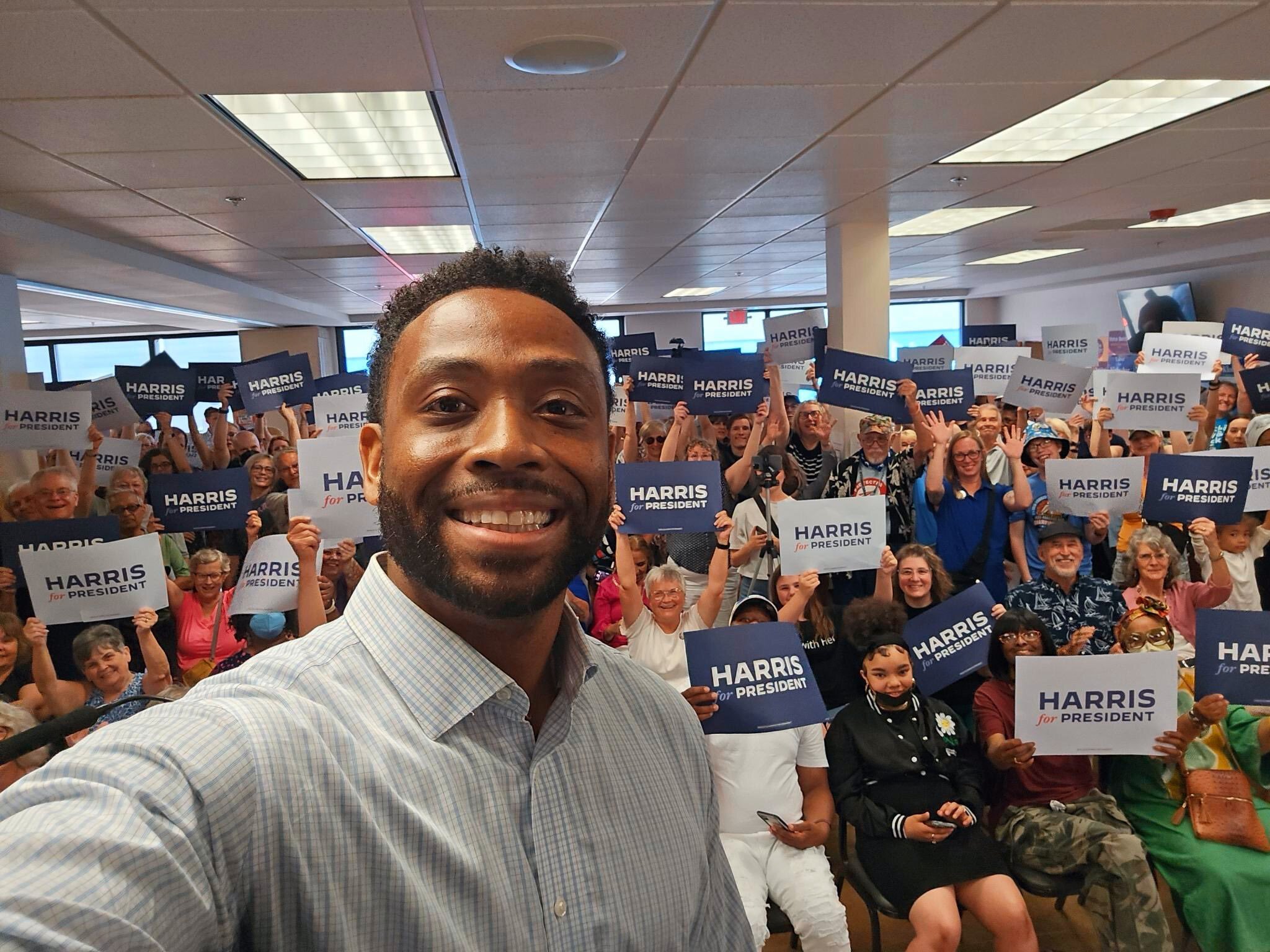 A room full of supporters hold rally signs while taking a selfie.