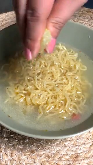 A close-up of a hand with pink nails squeezing lime over a bowl of instant noodles in broth, set on a woven mat.