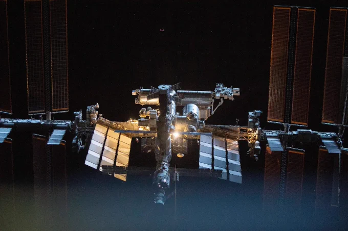 The International Space Station, as seen outside from a close distance. The station's solar panels frame the left and right sides of this photo, with its modules taking up much of its center. The background is dark and spotted with stars, with a blue glow fading up from the photo's bottom. Credit: NASA