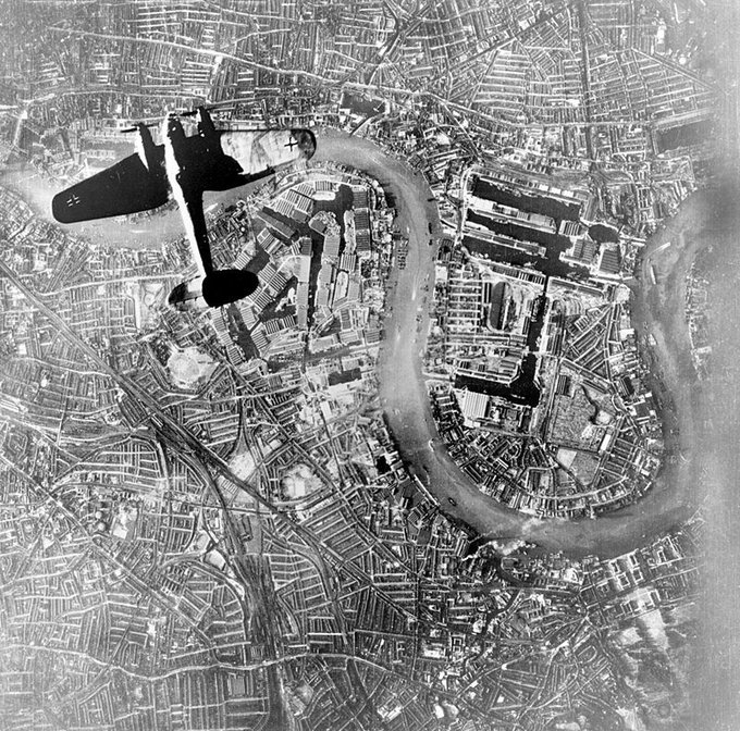A vintage black and white photo depicting an aerial view of a densely packed urban area with winding streets and river curves. Superimposed on the image is a silhouette of an old-fashioned airplane flying over the city, emphasizing the perspective of viewing from above.