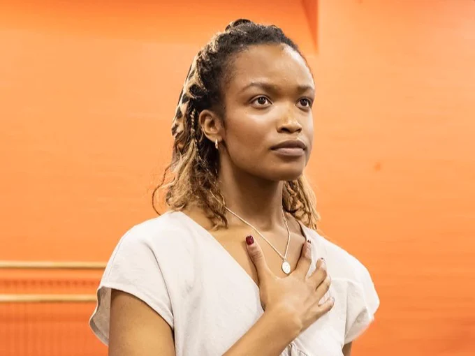 Person with braided hair wearing a white shirt, holding one hand over their heart, standing in front of an orange background.