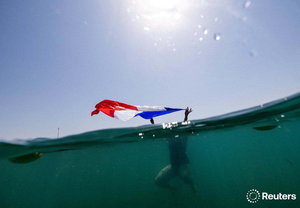 reuterspictures's tweet photo. A supporter of France waves the national flag as she swims before the women's windsurfing race at the #Paris2024 Olympics. More photos of the week: https://t.co/JwO0itCkLV 📷 Lisi Niesner https://t.co/MlpJUxNOso