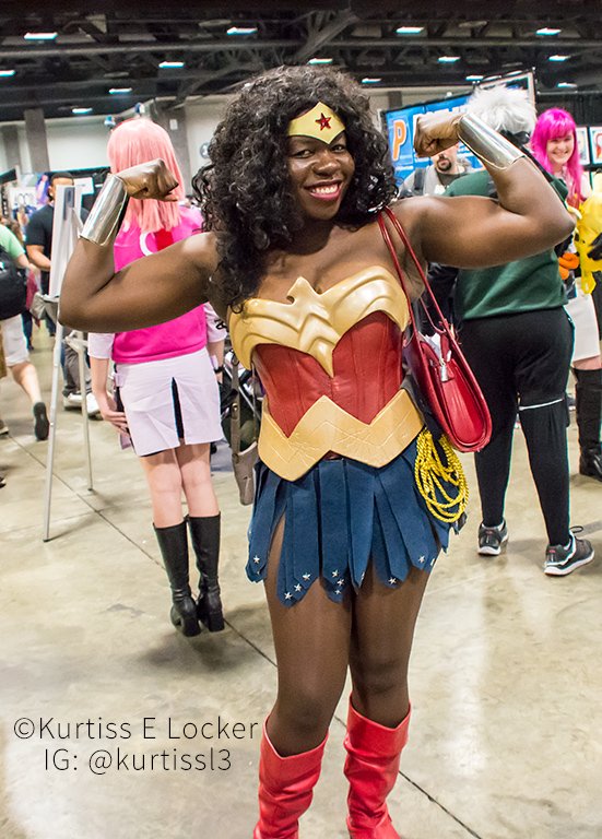 photo of a dark-skinned Black woman with long curly hair held back with a gold tiara on her forehead with a red star, wearing Wonder Woman armor with a gold W shaped breastplate with an eagle head, a gold W shaped belt over a red corset, blue divided skirts with silver stars and silver arm cuffs. she has a golden lasso on her hip, red boots and muscular arms. She has both arms flexed with a red purse on her shoulder, and is standing at a comic con. text on photo is the photographer credit "Kurtiss E Locker, IG:@kurtiss13"