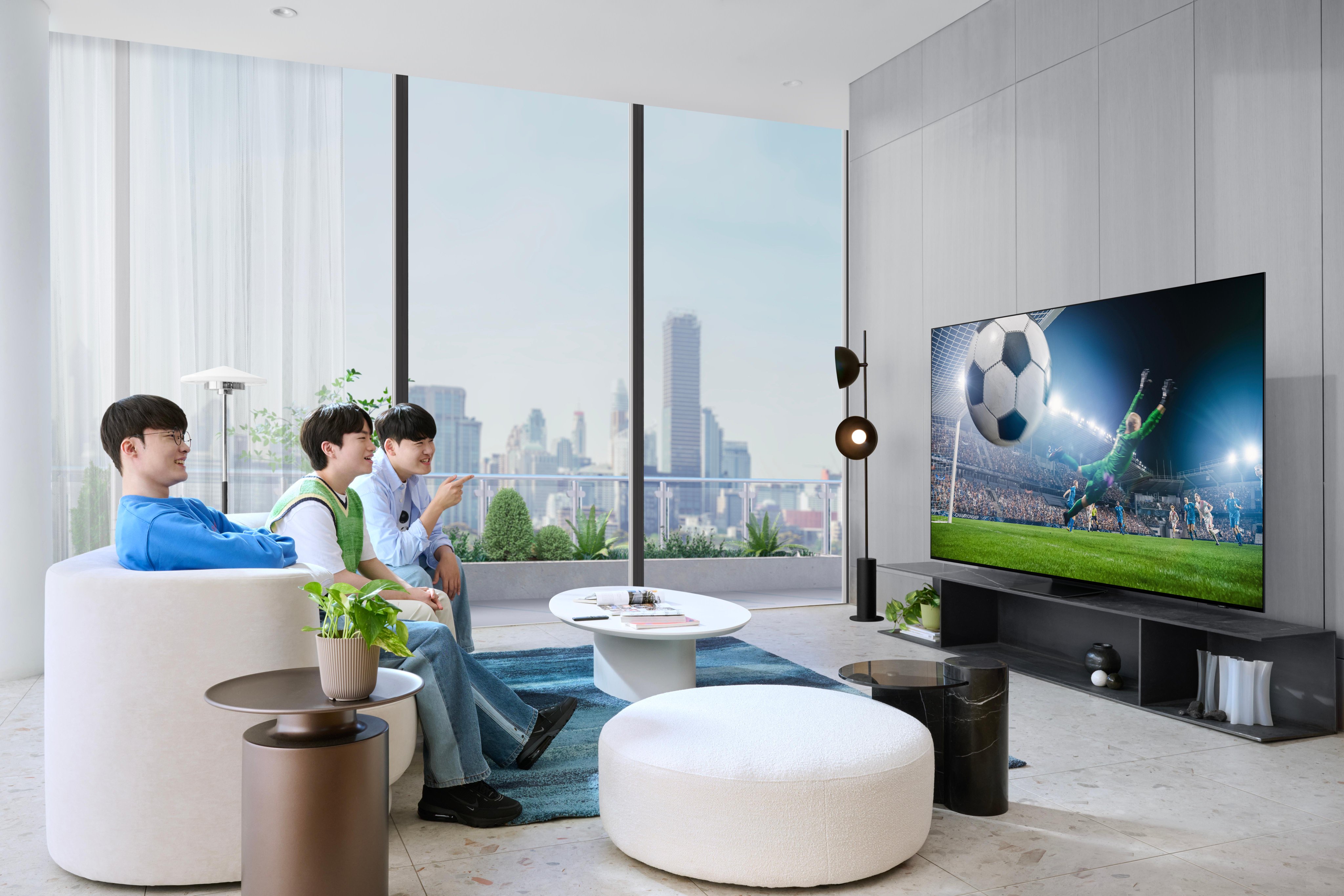 There is a room with a gray wall on one side and a large window on the other side that shows a building. The floor is made of marble, and there is a green carpet on the floor with a low white table on top of it. The Korean esports team T1 members are watching a soccer video on TV.