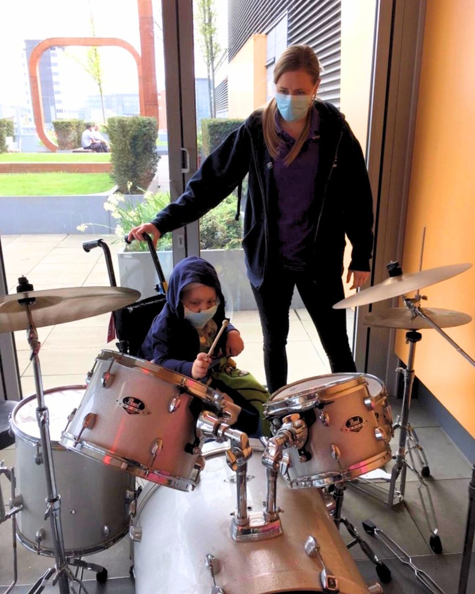 Play worker Alanna watches Jake play a drum set inside near a large window with an outdoor sculpture visible in the background.