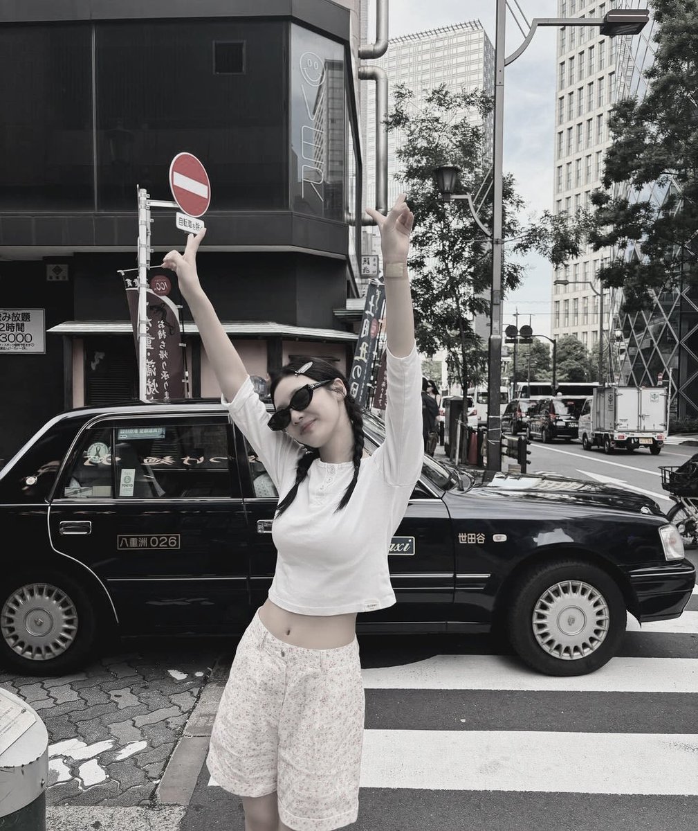 stands confidently at a crosswalk in an urban setting, blending casual chic with a playful touch. She wears a simple white, three-quarter sleeve top paired with high-waisted, floral print shorts that add a soft, feminine flair to her look.