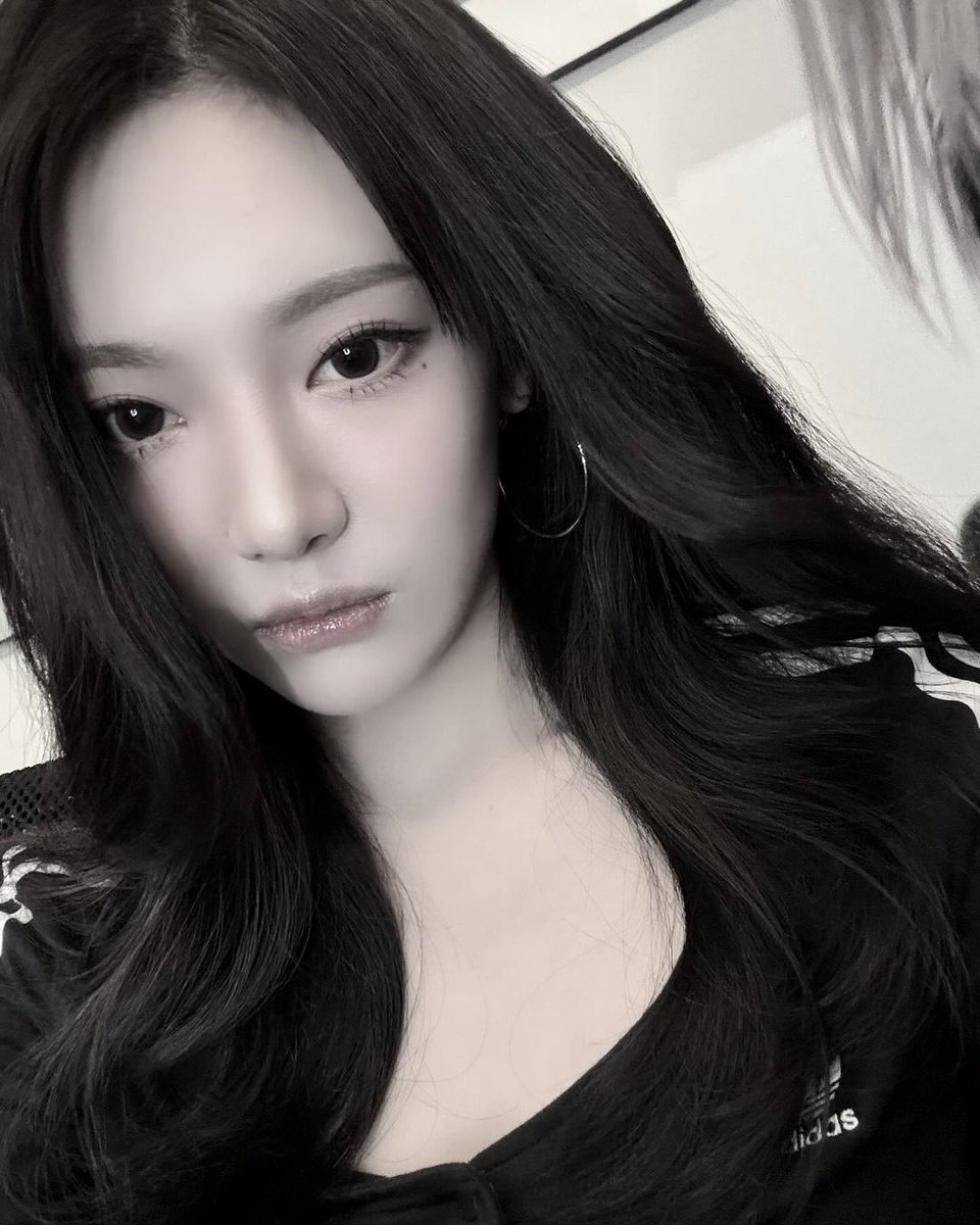 a young woman with long, dark hair looks directly into the camera with a serious expression. She has a soft pink blush on her cheeks and subtle makeup highlighting her eyes and lips. She's wearing a black top with an Adidas logo visible on her shoulder.