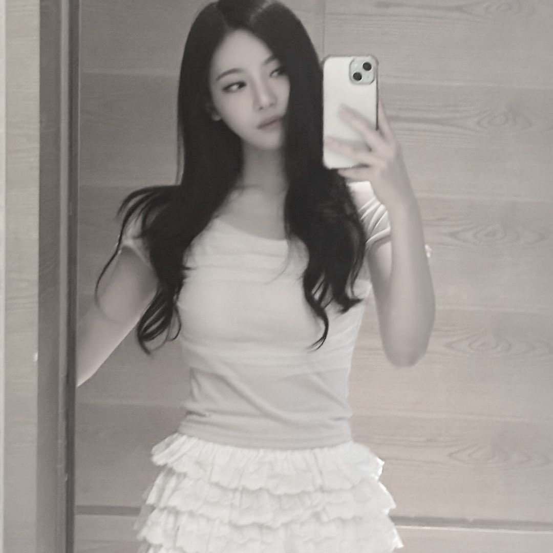 She has long, dark hair and is wearing a light pink top paired with a white, layered lace skirt. The background features a wooden panel, adding warmth to the image. The woman's pose is relaxed, with one hand holding her phone and the other resting by her side.