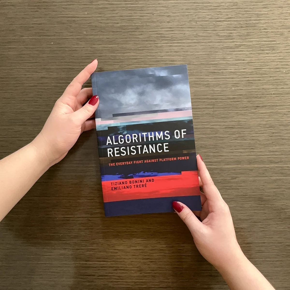 A person holds a book titled "Algorithms of Resistance: The Everyday Fight Against Platform Power" by Emiliano Treré and Tiziano Bonini, displaying the cover prominently against a wooden table background.