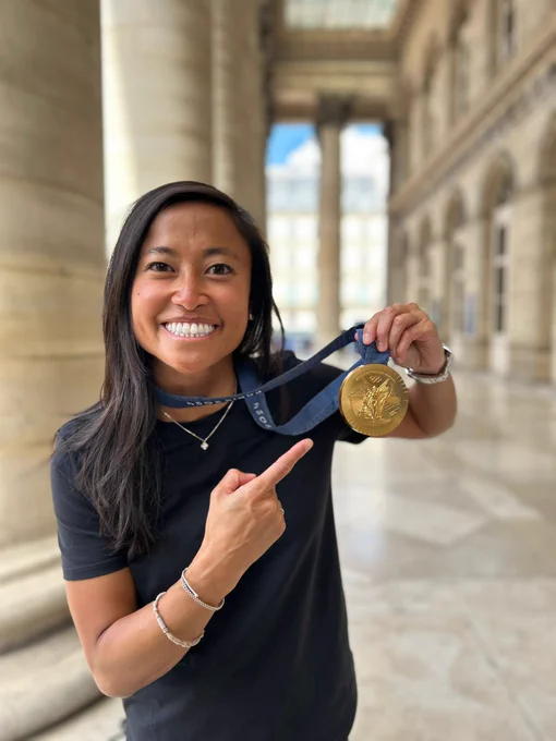 Person with long black hair poses pointing at an Olympic gold medal.
