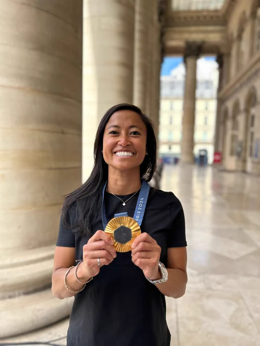 Person with long black hair poses with an Olympic gold medal. 