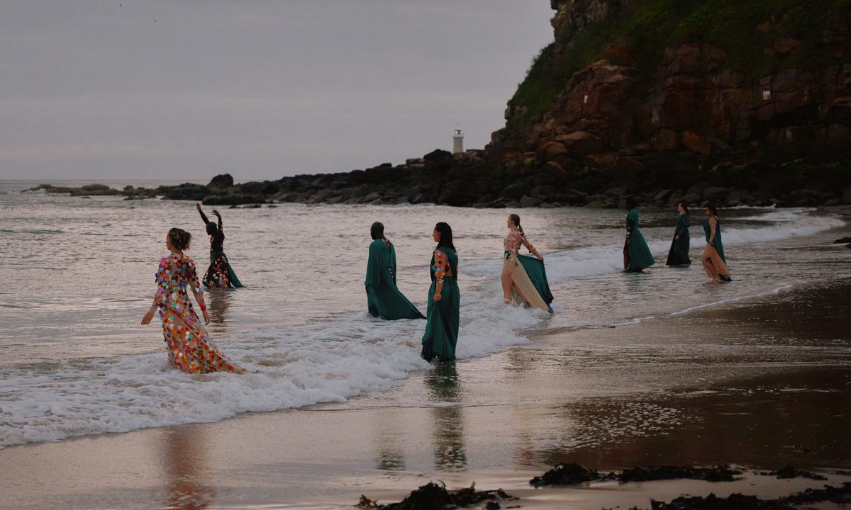 Into the sea, exploring experiences of grief.All performances of #GriefFloats at King Edward’s Bay, Tynemouth, will be #audiodescribed . 
The image shows the performers stepping into the water at sunrise, light glinting on the sequins of their robes. companyofothers.co.uk