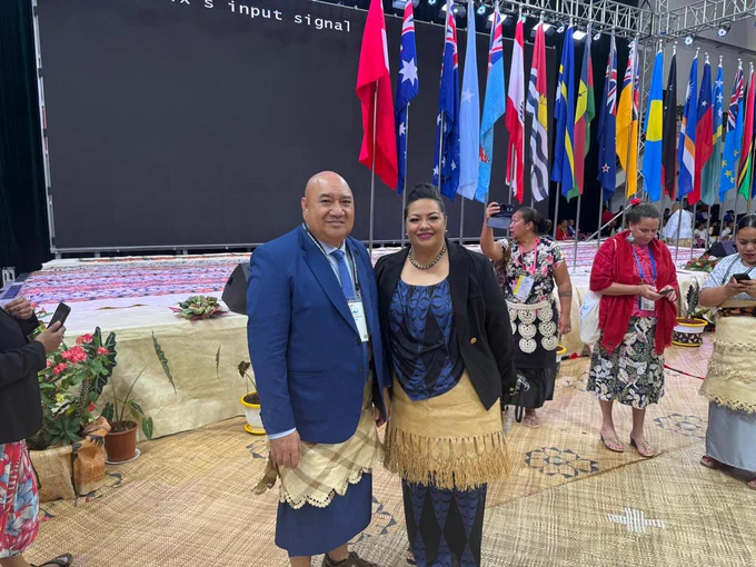 A man dressed in a blue suit stands next to a woman in a dress and blazer, both of them wear a woven ta’ovala. Behind them is a stage decorated with different flags and a few people on their phones 