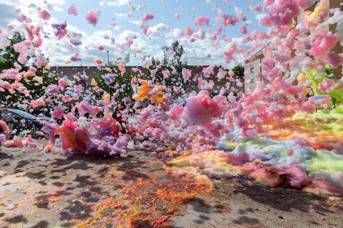 Colorful explosion of pink, orange, and green foam at a foam party under a sunny sky, with foam particles suspended in the air.