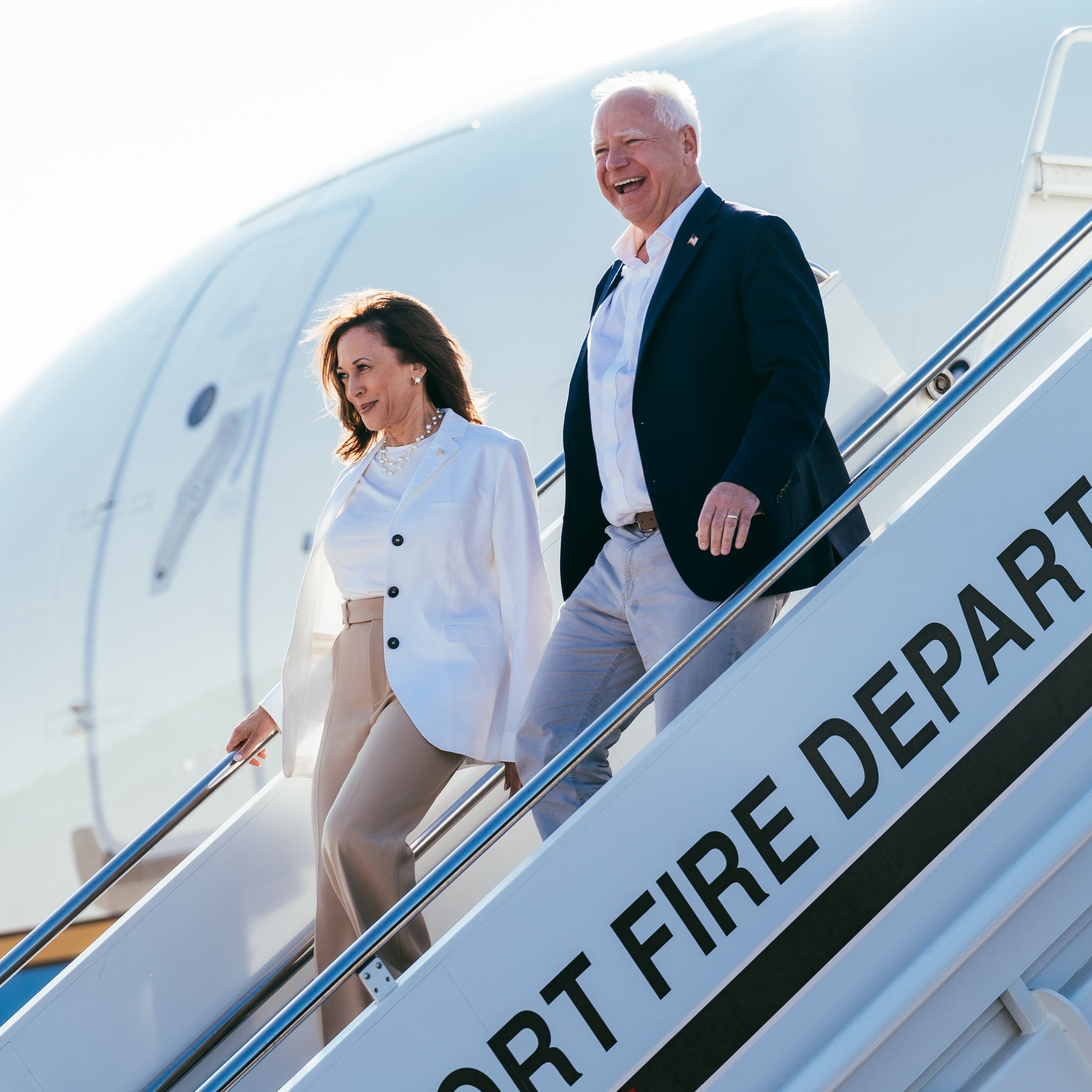 Vice President Kamala Harris and Governor Tim Walz descend the steps of a plane, both smiling and looking ahead. 