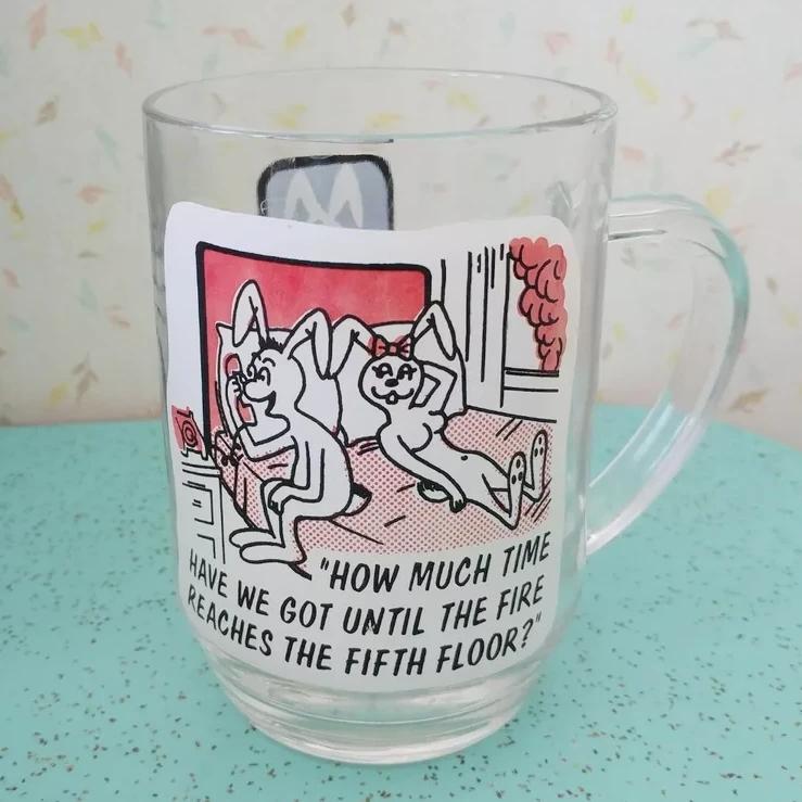 Glass mug with illustration of two rabbits in a hotel room, with a smoke plume outside. A femme rabbit is laying provocatively on the bed, while the masc-coded rabbit is on the phone asking "how much time have we got until the fire reaches the fifth floor?"