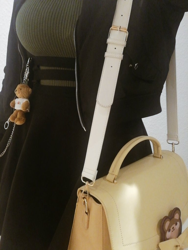 A closeup photo of a person standing in frame, wearing a black skirt and green pullover and a black jacket. The central point of the photo is a small bear plush hanging from the skirt and a large brown bag with a bear charm on it.