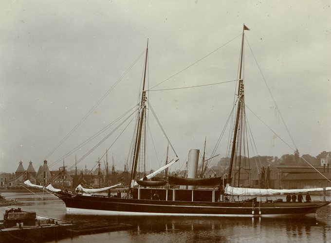 A vintage photograph depicts a docked steam-powered yacht with two tall masts, displaying a blend of sails and rigging. The vessel, featuring a prominent funnel, is moored in a calm harbour with an industrial backdrop. The background shows a collection of warehouses and buildings with distinctive pointed roofs. 