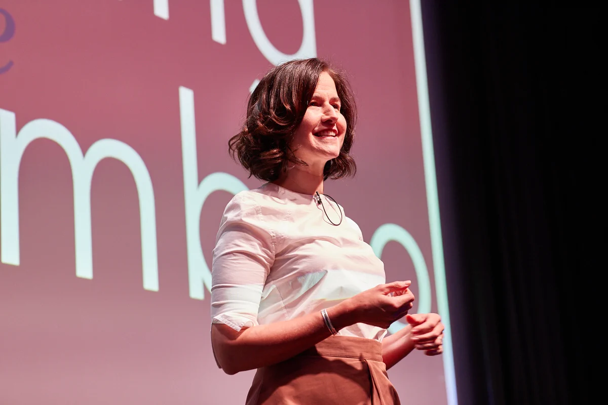 A woman with shoulder-length dark hair stands on stage, smiling and speaking. She wears a white blouse and high-waisted brown pants. The background features large, partially visible white text on a pinkish screen. The setting appears to be a formal presentation or talk.