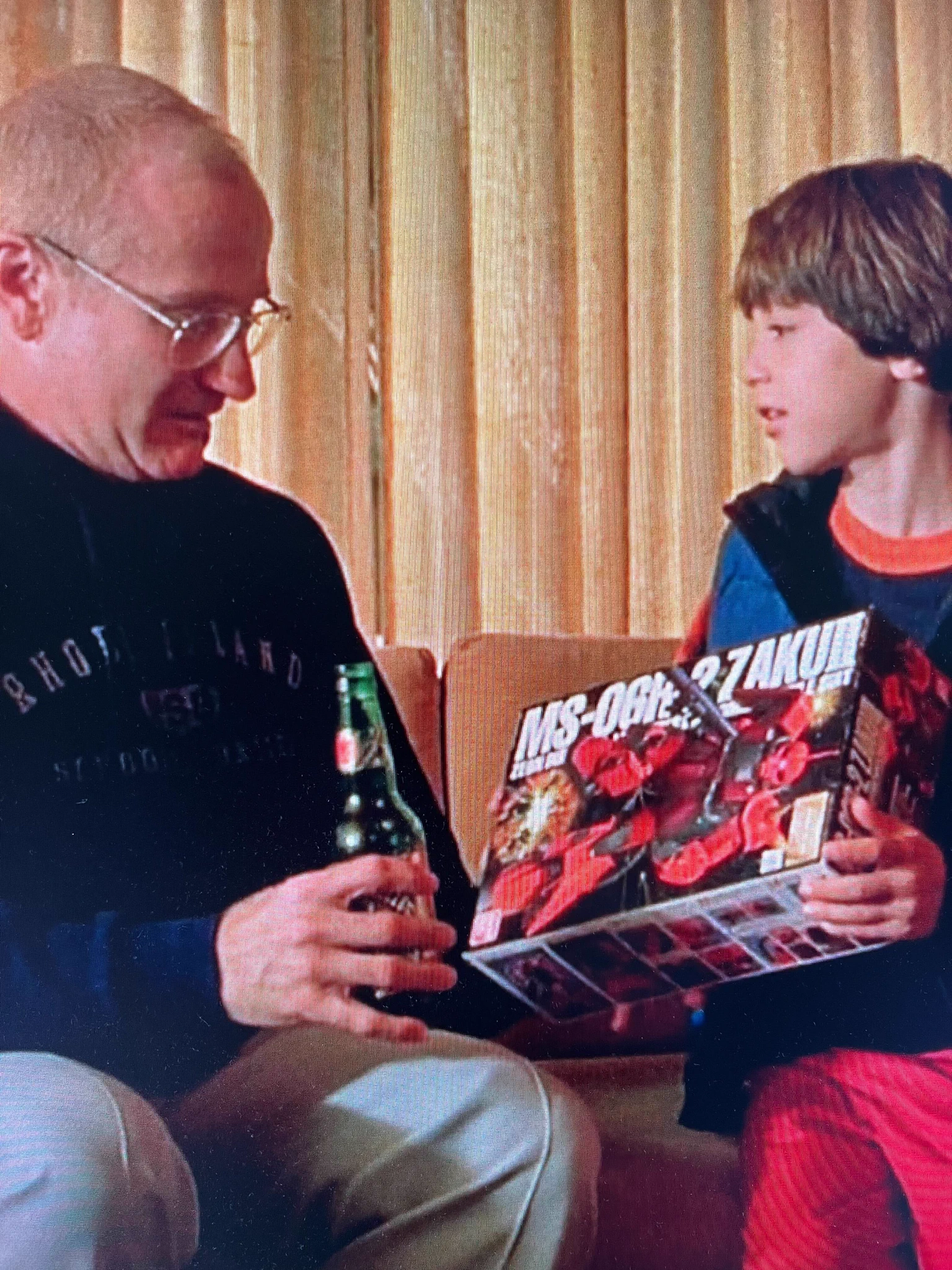 Robin Williams with blonde hair and a beer is being shown a box with a big red robot on it by a boy with short brown hair. The scene is about a half hour into One Hour Photo.