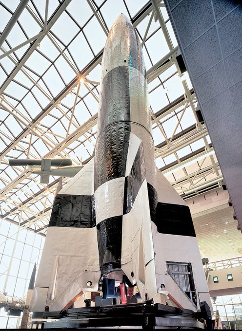 Display of a large rocket, featuring a black and white exterior, exhibited indoors under a geometric glass ceiling.
