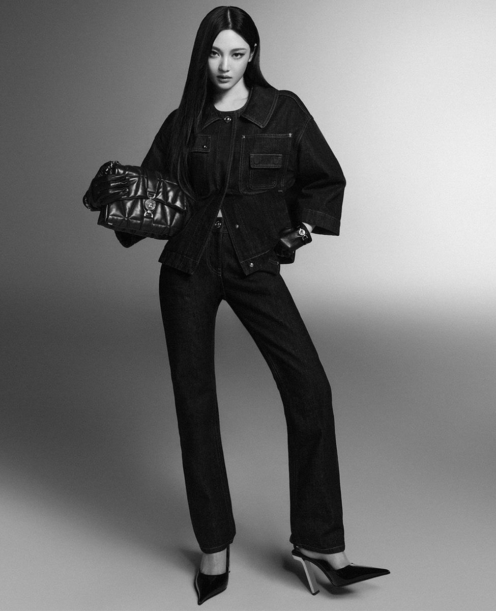 The subject is dressed in a textured, dark-toned coat, adorned with a "Versace" label on the sleeve, and is holding a sleek black leather handbag with a chain strap