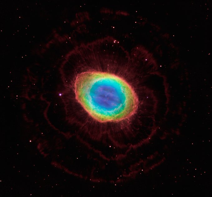 A round, prismatic nebula is on the black background of space, occupying about one-third of the field of view. The center is translucent blue gas with purple wisps. A tiny white star appears at center. Moving outward, the colors transition to teal, green, and yellow. The outermost portions are red, thicker, and very clumpy, and appear more oval, forming an outer ring. Tiny irregular knots of dense gas line the inner rim of the ring and point inward. Beyond the main nebula, faint dark red filaments form a rosette shape like petals on a flower. Dozens of pink stars are scattered across the field of view.