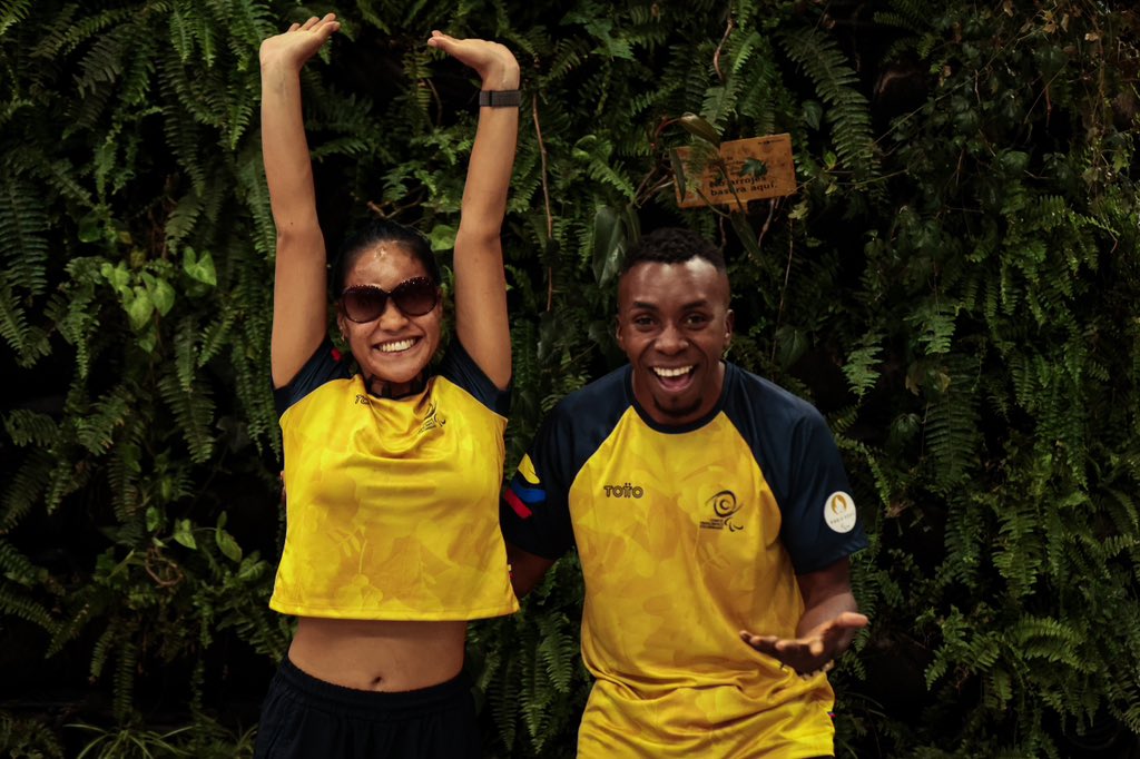 La imagen muestra a la Para atleta Angie Pabón junto a su guía de competencia, Luis Arizala, posando con alegría. Ambos visten camisetas amarillas de la delegación nacional, mostrando su orgullo y entusiasmo, de fondo se observan plantas.