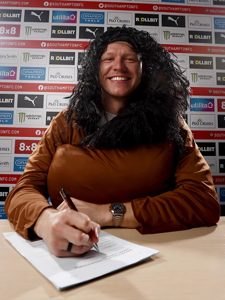 An image of Aaron Ramsdale, wearing a Hagrid fancy dress costume, smiling at the camera while signing his Saints contract in front of a sponsors' back drop.