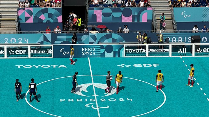 Foto del juego entre Colombia y Japón durante los Juegos Paralímpicos París 2024.  El campo es de color azul y está marcado con el logo de “París 2024” en letras blanca. El equipo nacional usa camiseta amarilla con pantaloneta blanca, mientras que el rival usa color azul. En el fondo, se ve la gradería con muchos espectadores.