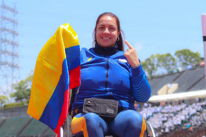 Erica Castaño, con una sonrisa en la cara, sostiene la bandera de Colombia y con el dedo indice de su mano izquierda marca el número uno. Lleva una sudadera azul. 