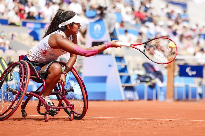 Foto de Angélica, tenista en silla de ruedas durante un juego en cancha de arcilla. Extiende la raqueta de color blanco con rojo con su brazo derecho para golpear la pelota de color verde encendido. En el fondo, se pueden ver espectadores.