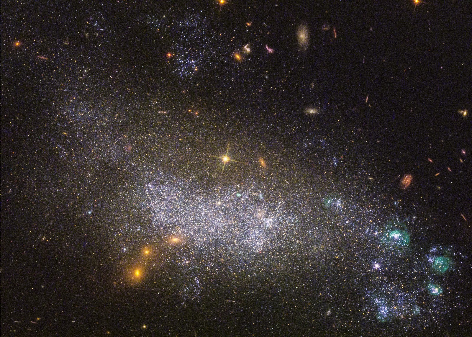 Irregular galaxy UGC 5340: A dense, but diffuse bean-shaped star field occupies the center and lower left portion of the frame. Stars in the outer portion of the dwarf galaxy appear yellow, while the brighter stars near the galaxy’s core are blueish. At lower right are additional clumps of bright, blue stars, as well as several small, green bubbles of gas. Several dozen background galaxies of various shapes and sizes are scattered across the image. The background of space is black.