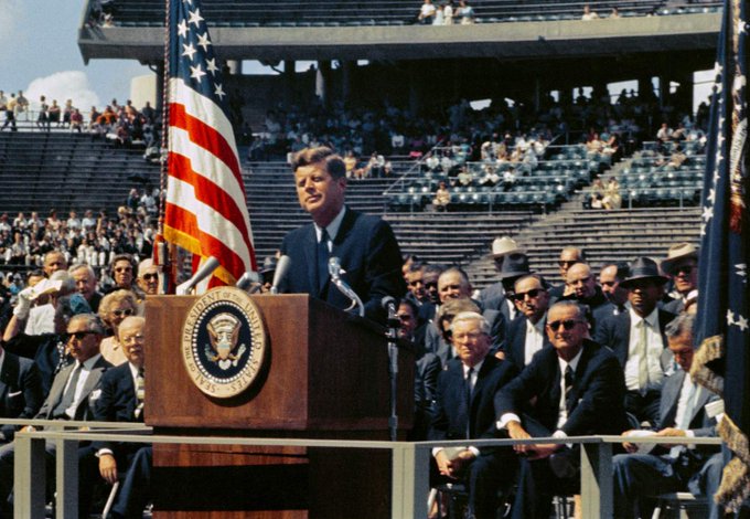 President John F. Kennedy delivering a speech at a podium with the Presidential Seal, an American flag in the background, and a crowd of spectators at an outdoor event.