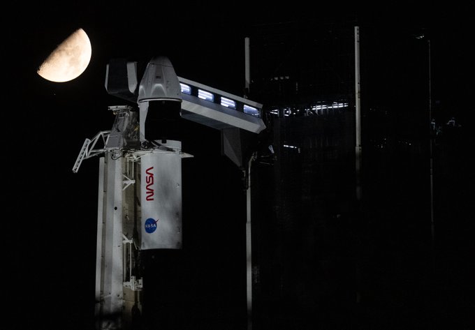A half-full Moon rises above a SpaceX Falcon 9 rocket at NASA's Kennedy Space Center. The white rocket, with an access arm attached to the Dragon spacecraft at top, takes up most of the image, with the Moon above and to the left of it. Credit: NASA/Joel Kowsky