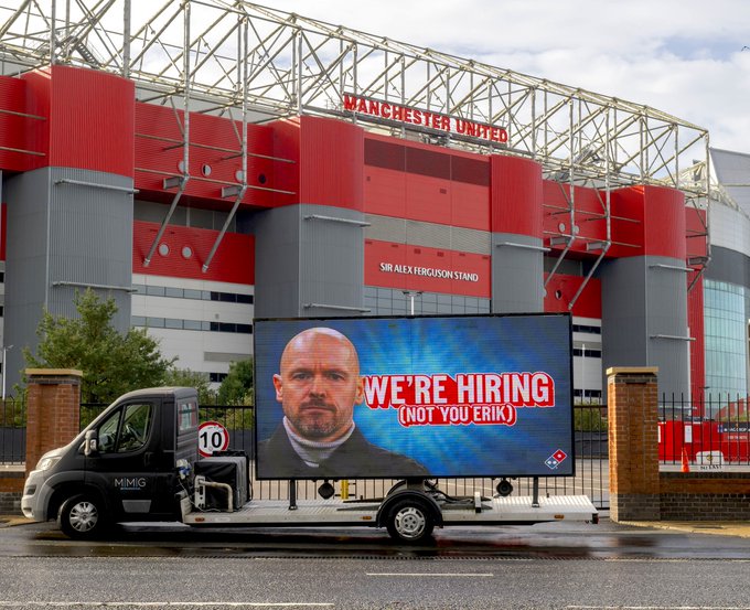 An advertising truck parked outside Manchester United's football stadium, featuring a digital billboard with a message saying 'WE'RE HIRING (NOT YOU ERIK)' and an image of a stern-looking Erik ten Hag.