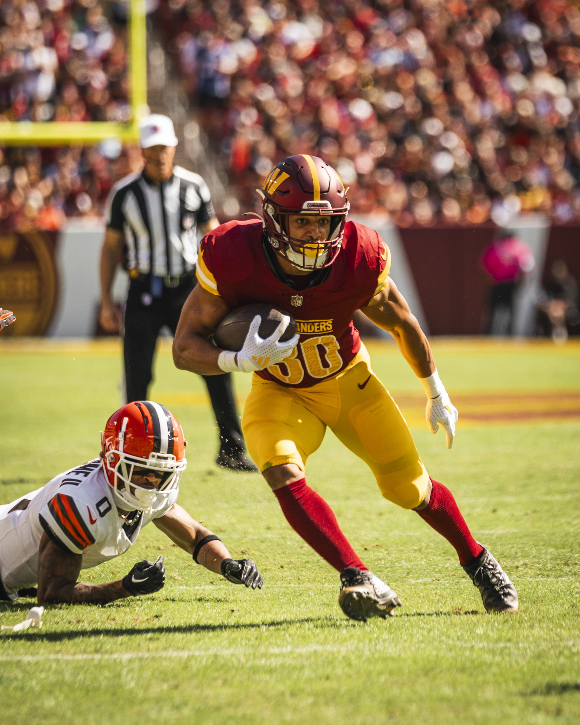 Photo of Austin Ekeler running. He is holding a football in his right hand. He is wearing a burgundy helmet, burgundy jersey, gold pants, white football gloves and burgundy socks.