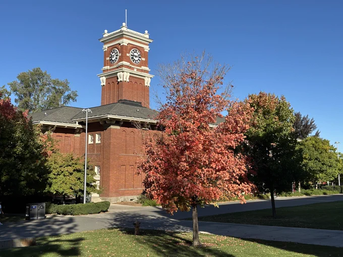 Color outdoor photo during daytime. Orange and red leaves are on a tree in the foreground with blue sky above. A red brick building is in the background with a clock tower. 