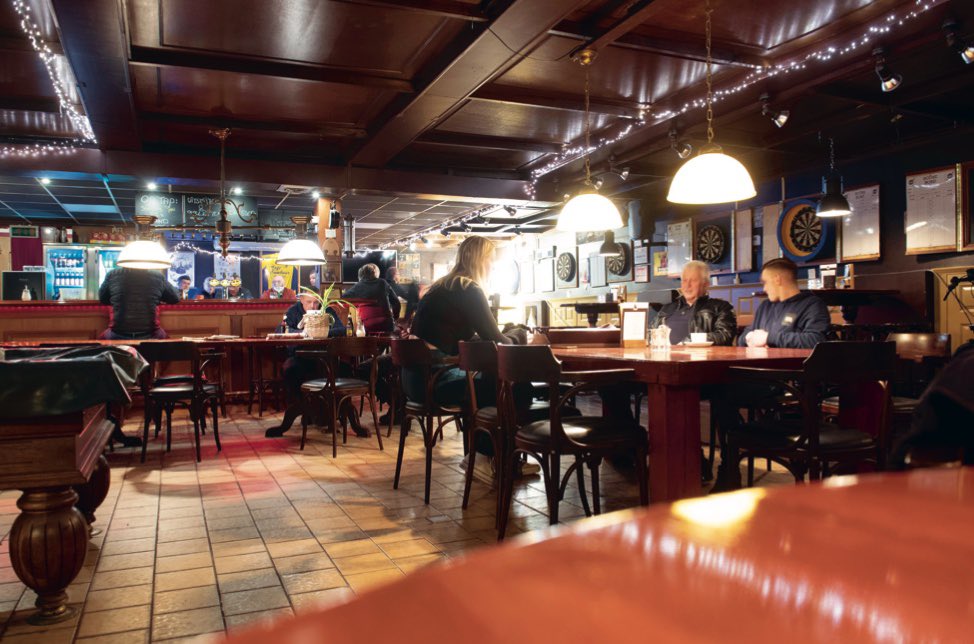 Staantribune's tweet photo. Recht tegenover de Zuidtribune van het oude Cambuurstadion staat zonder twijfel het beste supporterscafé van Nederland: Café Cambuur, in de volksmond beter bekend als de Cambuurbar.

Op een wedstrijddag stonden de rode fietsenrekken al vroeg vol met tweewielers. Buiten drommen groepen supporters samen en binnen in het donkere café is het altijd druk. Des te belangijker de wedstrijd, des te vroeger lijkt men hier de zenuwen weg te willen drinken. De aankleding van het in 1969 geopende café alleen al is uniek. Bij de ingang word je verwelkomd door een beeld van een clown, terwijl er verderop een oude, hou- ten indiaan te vinden is. Aan de muren hangen willekeurige memorabilia en foto’s van de club, de wijk en de stad. Het is een heerlijke ratjetoe van voorwerpen, die worden verlicht door de altijd aanwezige kerstverlichting. In het oude rookhok dient een oude typmachine, die half onder een piano is weggestopt, als extra stoel aan een lange tafel. Aan de overkant hiervan zit altijd wel iemand achter de gokkast.

Op wedstrijddagen was het concept simpel. Je wurmt je langs de menigte die buiten staat naar binnen, slaat rechtsaf en haalt bij een tafel in een hoek van het café blauwe muntjes. De muntjes worden bij de bar omgezet in flesjes Hertog Jan, of voor een enkeling cola Berenburg. Op de pooltafels staan lege kratten waarin je de lege bierflesjes kunt retourneren. En wie echt dorst heeft, haalt gewoon een volledig volle krat bij de bar.

Meer over De Cambuurbar  en het Cambuurstadion in ons boek ‘Estadio deel 4️⃣’, die geheel in het teken staat van het oude stadion aan het Cambuurplein. 

Bestel, zolang de voorraad nog strekt, via ⬇️
https://t.co/MY7ZT0p31u

🚨 NB: nog slechts 24 exemplaren!