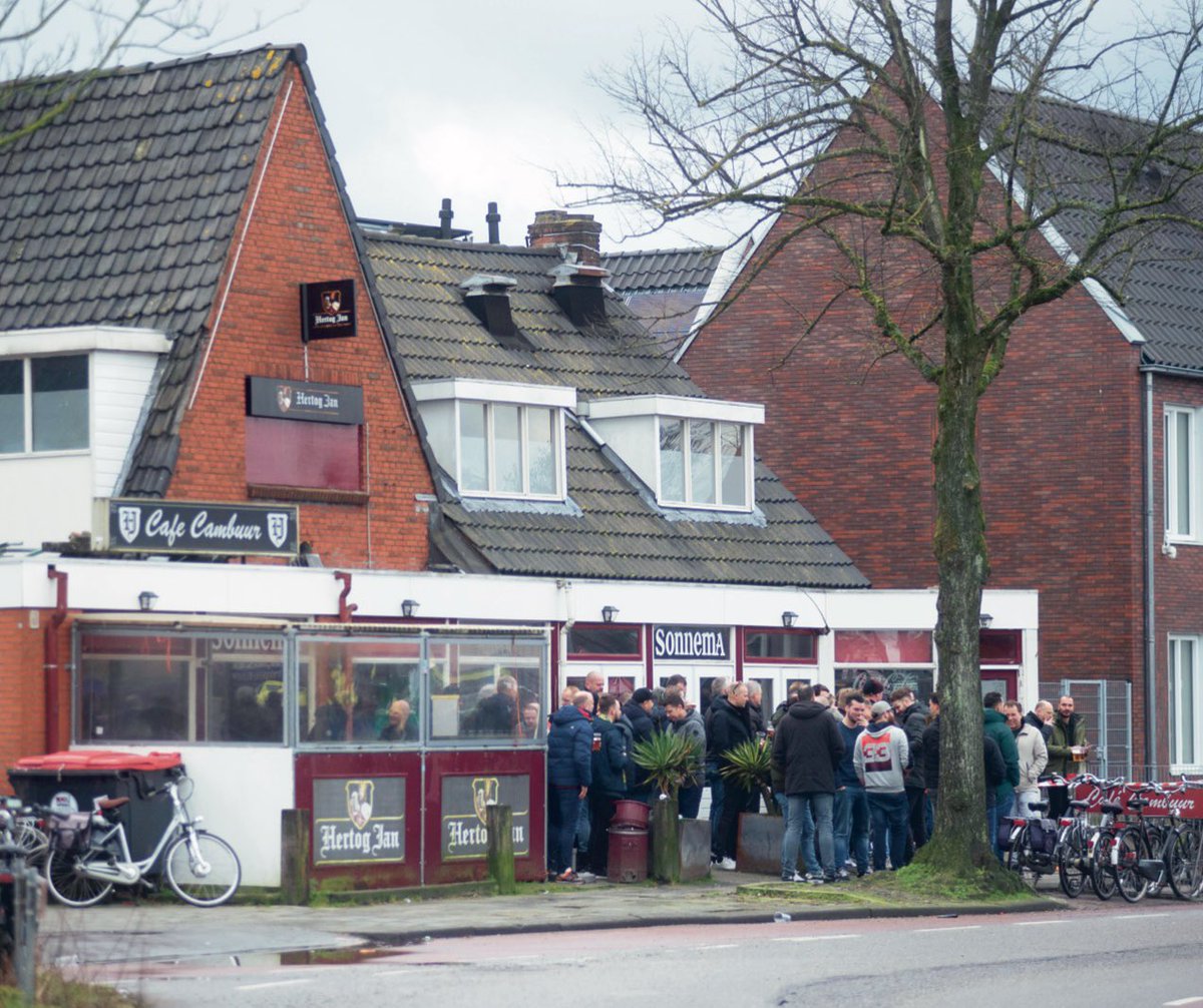 Staantribune's tweet photo. Recht tegenover de Zuidtribune van het oude Cambuurstadion staat zonder twijfel het beste supporterscafé van Nederland: Café Cambuur, in de volksmond beter bekend als de Cambuurbar.

Op een wedstrijddag stonden de rode fietsenrekken al vroeg vol met tweewielers. Buiten drommen groepen supporters samen en binnen in het donkere café is het altijd druk. Des te belangijker de wedstrijd, des te vroeger lijkt men hier de zenuwen weg te willen drinken. De aankleding van het in 1969 geopende café alleen al is uniek. Bij de ingang word je verwelkomd door een beeld van een clown, terwijl er verderop een oude, hou- ten indiaan te vinden is. Aan de muren hangen willekeurige memorabilia en foto’s van de club, de wijk en de stad. Het is een heerlijke ratjetoe van voorwerpen, die worden verlicht door de altijd aanwezige kerstverlichting. In het oude rookhok dient een oude typmachine, die half onder een piano is weggestopt, als extra stoel aan een lange tafel. Aan de overkant hiervan zit altijd wel iemand achter de gokkast.

Op wedstrijddagen was het concept simpel. Je wurmt je langs de menigte die buiten staat naar binnen, slaat rechtsaf en haalt bij een tafel in een hoek van het café blauwe muntjes. De muntjes worden bij de bar omgezet in flesjes Hertog Jan, of voor een enkeling cola Berenburg. Op de pooltafels staan lege kratten waarin je de lege bierflesjes kunt retourneren. En wie echt dorst heeft, haalt gewoon een volledig volle krat bij de bar.

Meer over De Cambuurbar  en het Cambuurstadion in ons boek ‘Estadio deel 4️⃣’, die geheel in het teken staat van het oude stadion aan het Cambuurplein. 

Bestel, zolang de voorraad nog strekt, via ⬇️
https://t.co/MY7ZT0p31u

🚨 NB: nog slechts 24 exemplaren!