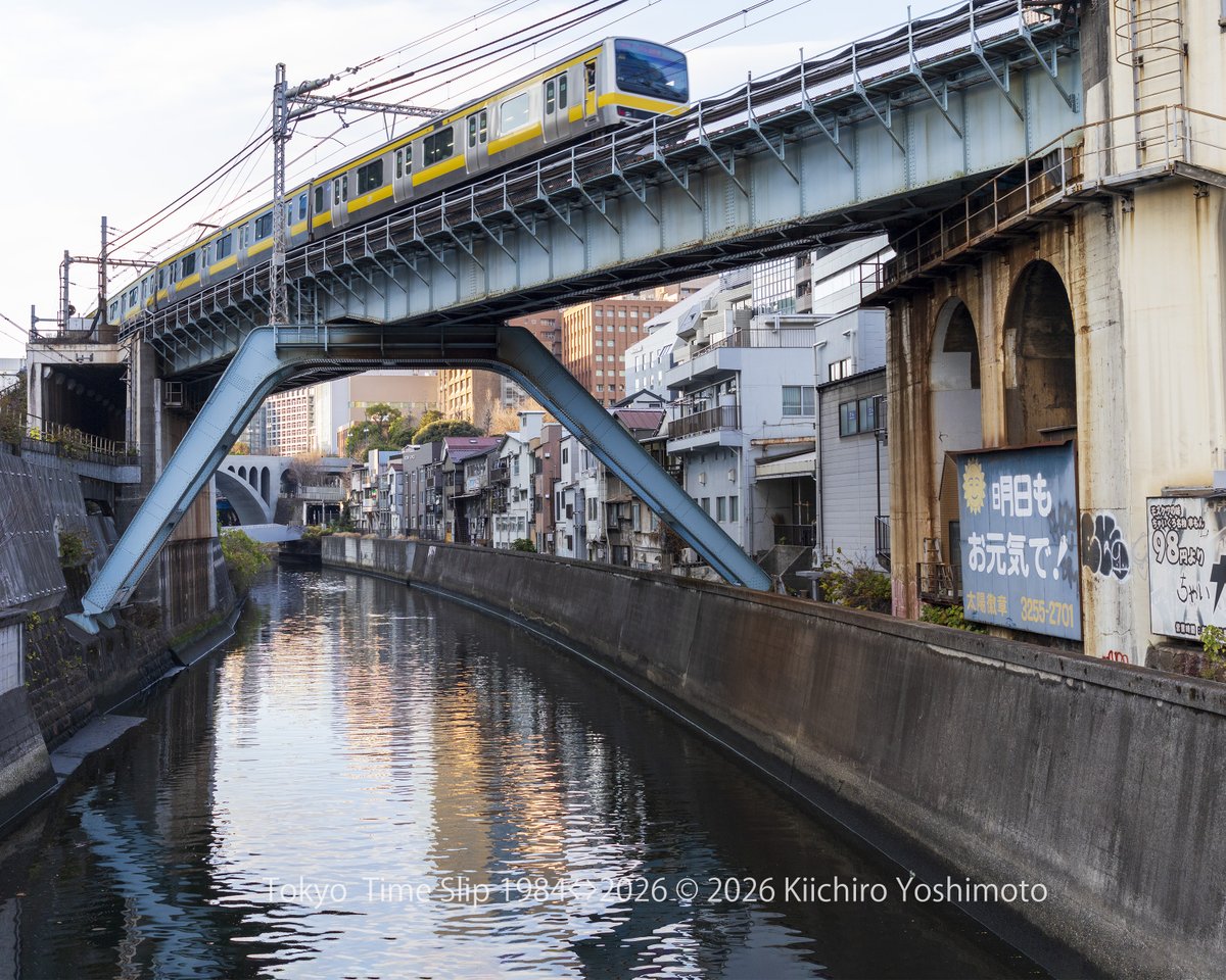 秋葉原・神田川橋梁 1984⇔2021⇔2026
神田川に架かる総武本線の鉄道橋。
1984年の写真では、右手の看板に「今日もお元気で！」の文字。2021年には「明日もお元気で！」に変わり、時代の移ろいを感じさせます。
奥に見えるのは聖橋。川沿いの風景は、今も昔も変わらぬ静けさをたたえています。