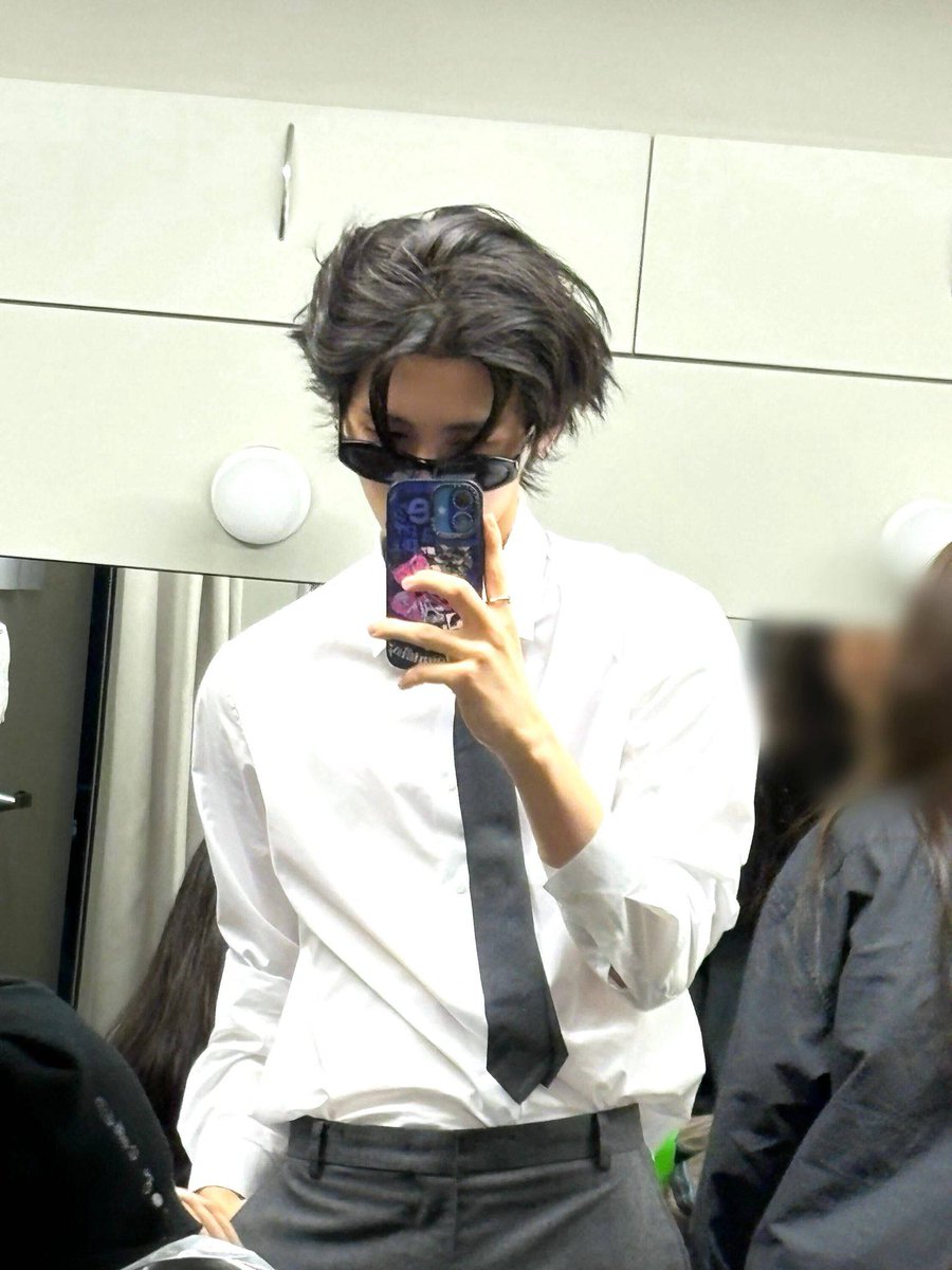 Untamed black locks fall deliberately, disrupting symmetry above a poised frame. A crisp white shirt contrasts a loosely set tie, the harmony exact and never rigid. Sharp tailoring blends with subtle disarray, creating a statement defined by contrast instead of extravagance.