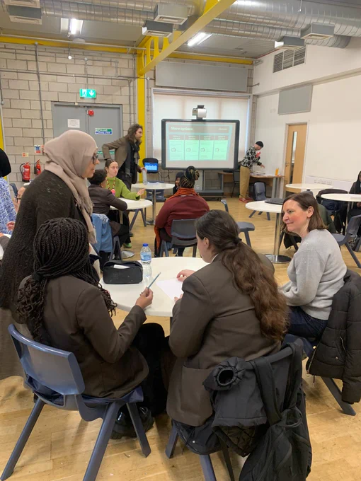 Vicky (right) engaged in group discussion with Addey and Stanhope students in a classroom. There is a presentation displayed on a screen at the front.