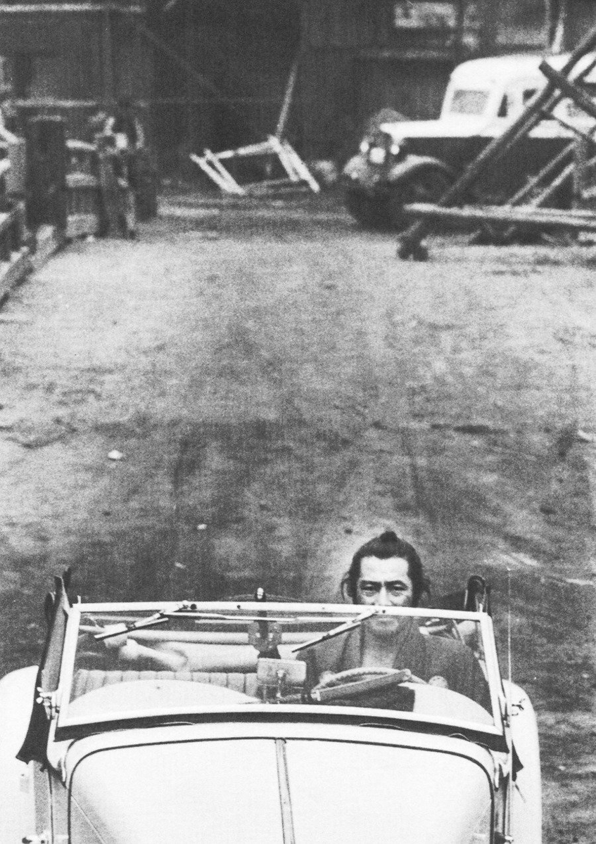 Toshiro Mifune driving on the set of Yojimbo, with his sword resting on the dashboard.