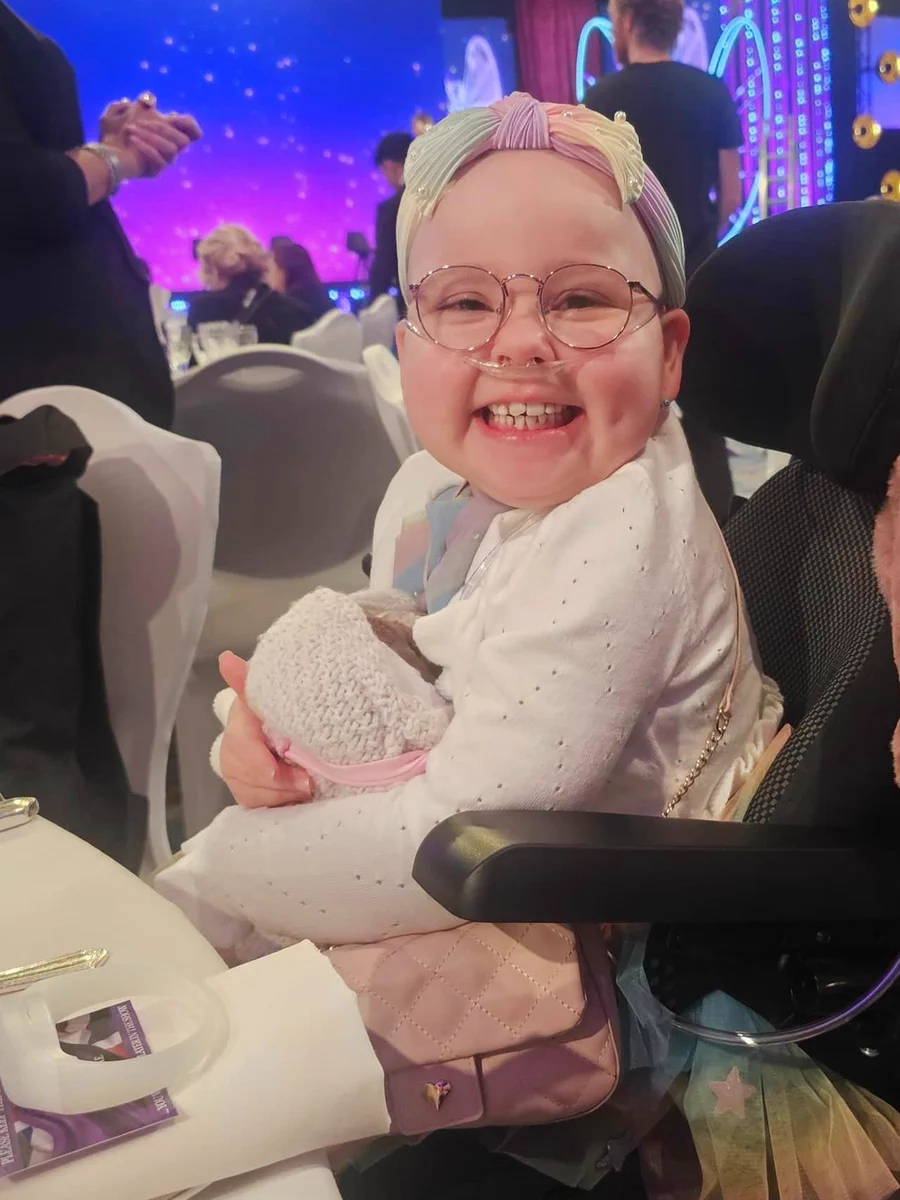 A young girl is sitting in a wheelchair wearing a bright coloured headband and beaming at the camera. 