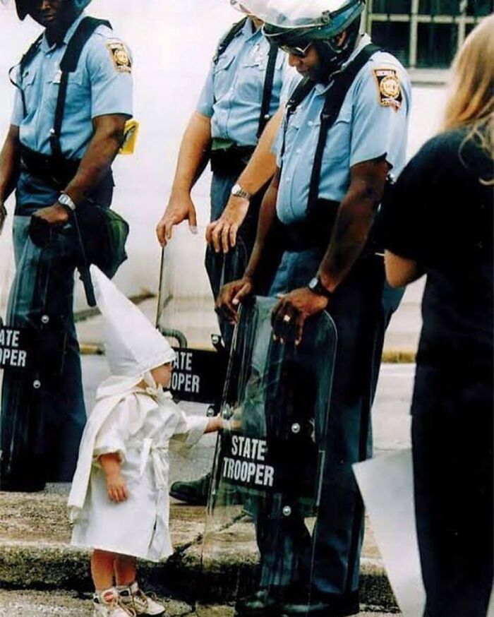12. "Nadie nace racista" Fotografía de Todd Robertson. Un niño vestido con una túnica y capucha del KKK, toca curiosamente el escudo de un policía durante un mitin del KKK | Gainesville, Georgia, 1992