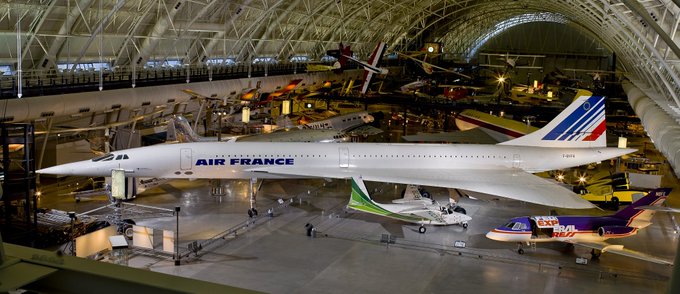 An Air France Concorde supersonic passenger jet is displayed prominently in a large hangar with various other aircraft, including a Boeing 747 and smaller jets, visible in the background.
